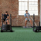 Two men performing battle rope exercises on green turf in industrial gym space with brick walls