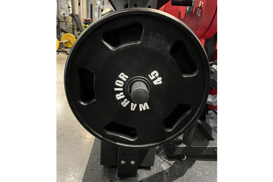 Black Olympic weight plate with grip handles and white text reading "Warrior" and "25 lb", displayed in gym setting
