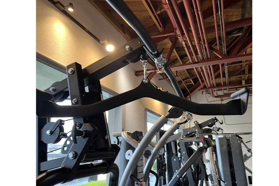 Black and silver home gym equipment with curved handlebars, leg press attachment, and cable pulley system in gym setting