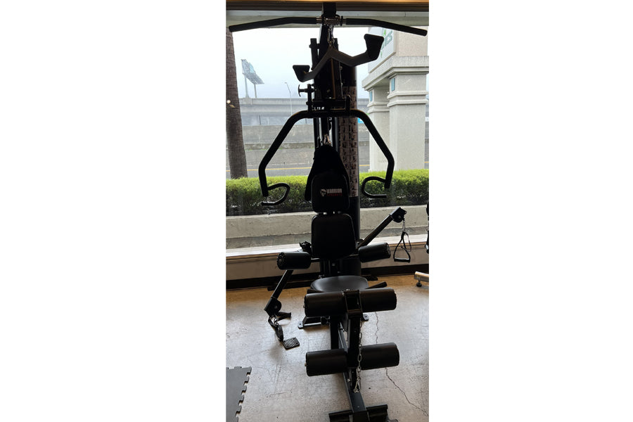 Black home gym equipment with curved handles, seat, and leg press attachment, photographed indoors by window