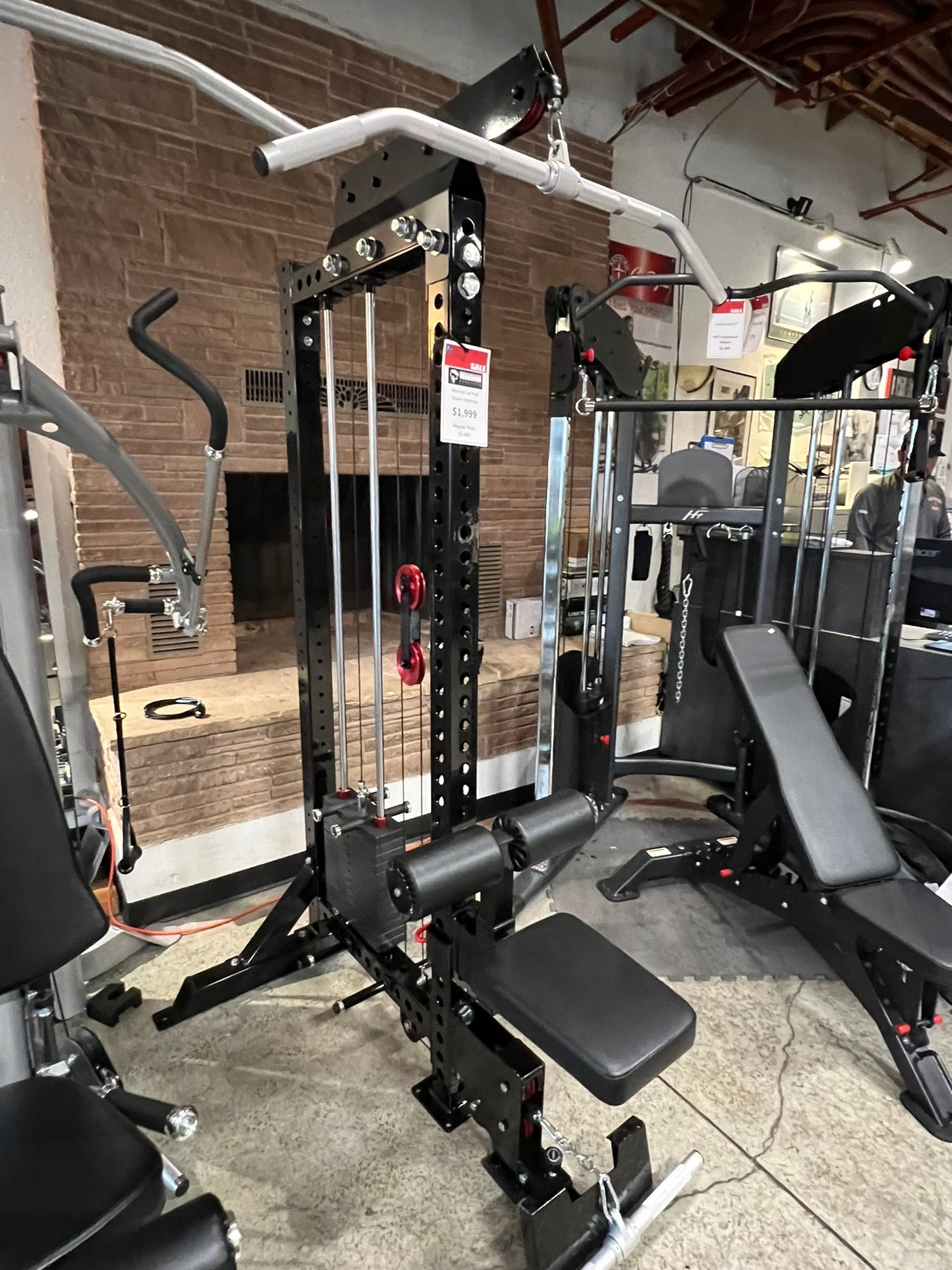 Black steel lat pulldown and low-row home gym system with white overhead bar, red weight pins, and black padded seat, display