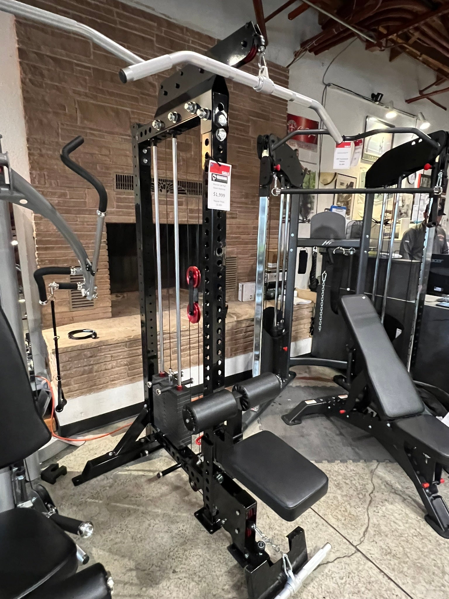Black steel lat pulldown and low-row home gym system with white overhead bar, red weight pins, and black padded seat, display