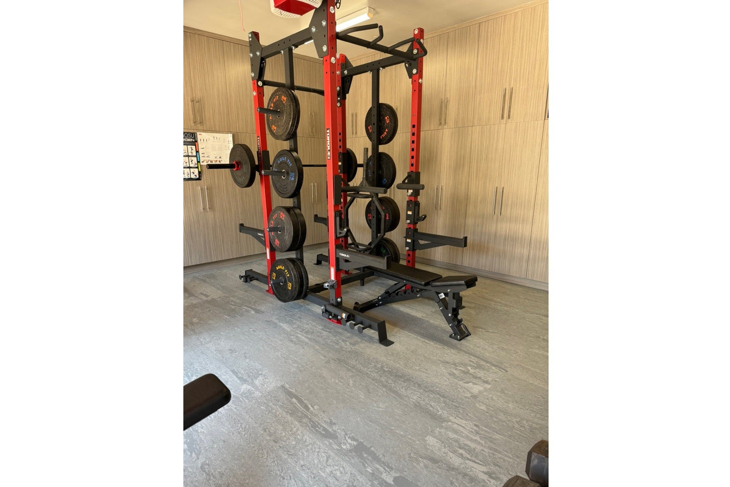 Red and black power rack with weight plates and barrels in gym setting on grey interlocking tile flooring
