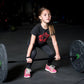 Young girl in black t-shirt with red graphic lifting barbell with green and black weight plates in gym