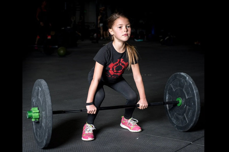 Young girl in black t-shirt with red graphic lifting barbell with green and black weight plates in gym
