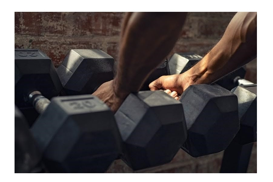 Person's hand gripping black rubber hexagonal dumbbell from a set, displayed on gym floor with brick wall background
