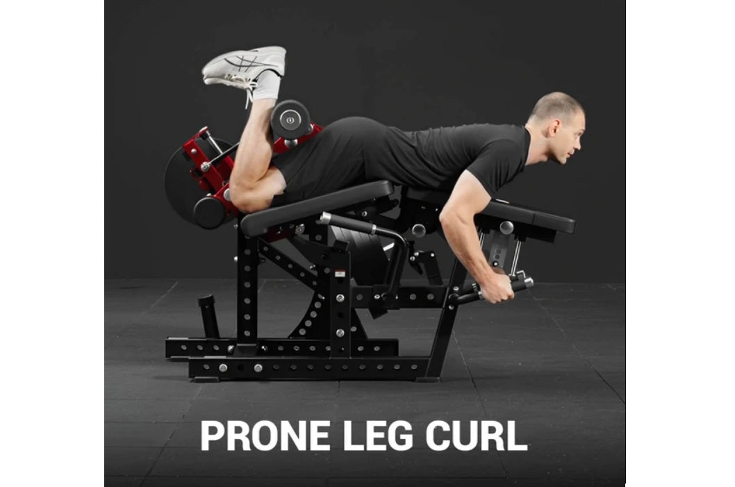 Man performing prone leg curl on black plate-loaded Warrior Signature machine, holding red dumbbells, wearing white sneakers 