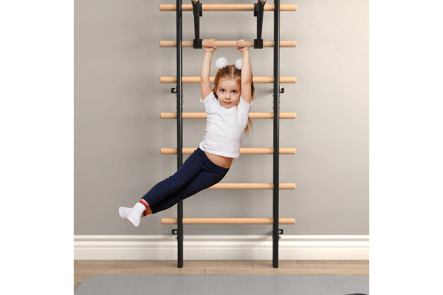 Young child wearing white t-shirt and navy leggings hanging from wooden ladder bars mounted on wall