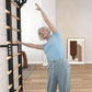 Senior woman in light blue t-shirt and gray pants exercising on black and wood wall-mounted ladder bars in home gym setting