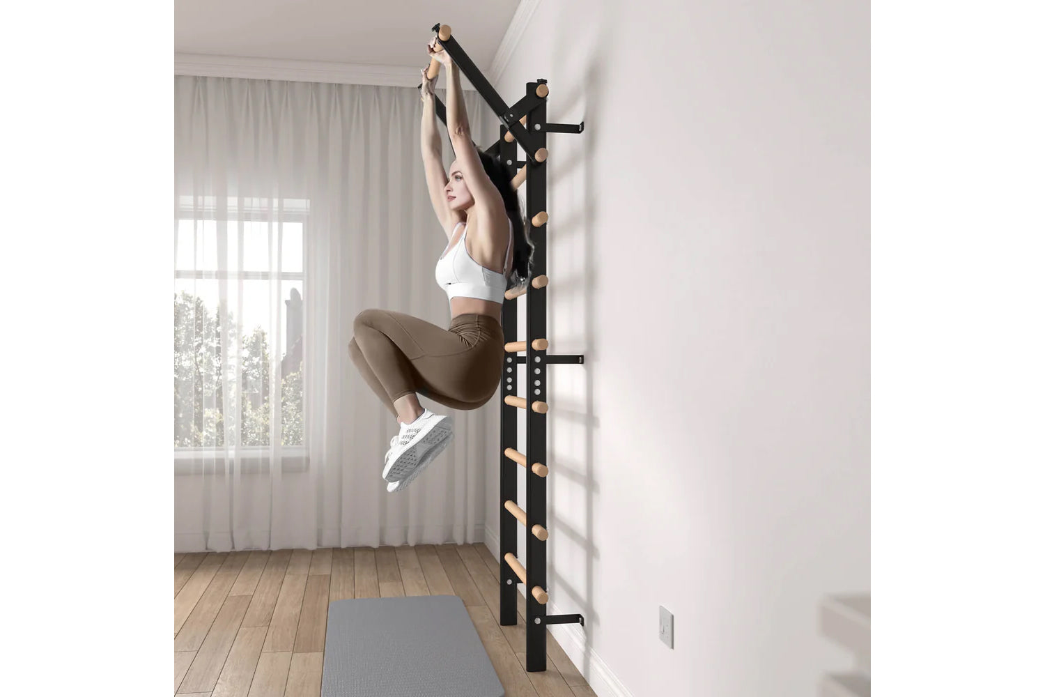 Woman wearing white sports bra and tan leggings using black wall-mounted ladder bars with wooden rungs in bright home gym