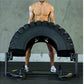 Shirtless man performing tire flip exercise with black oversized tire on metal frame in gym setting