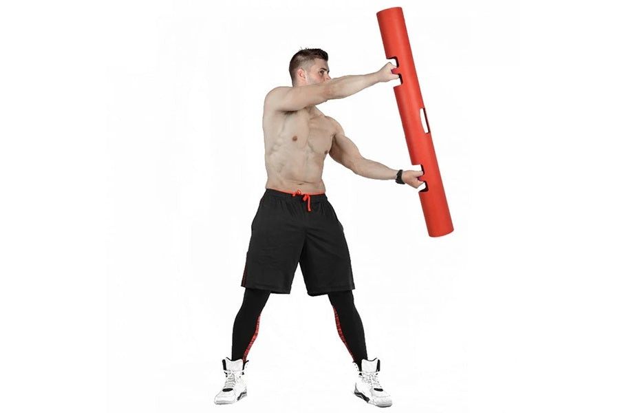 Shirtless man holding red foam training tube during workout, wearing black shorts with red trim and white sneakers