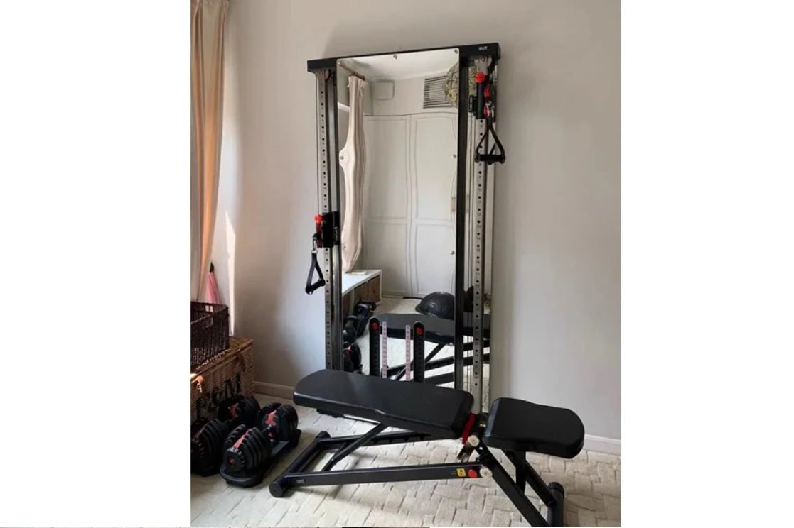 Black steel wall-mounted cable pulley gym system with dual weight stacks, mirror, and adjustable bench in home fitness space