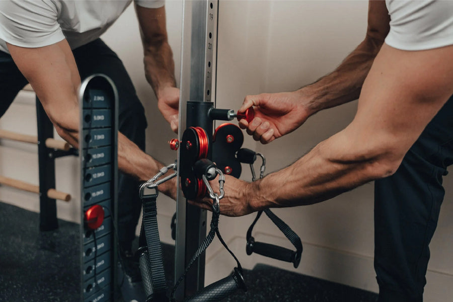 Two people assembling black and red cable pulley system mounted on wall, showing dual stack weights and carabiners