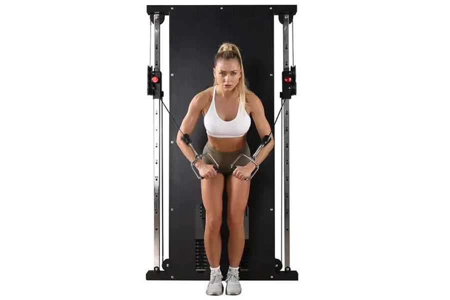 Woman using black and silver cable pulley home gym system with dual weight stacks, wearing white sports bra and olive shorts