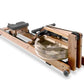 WaterRower oak rowing machine with natural wood frame, black padded seat, and water resistance chamber, shown fully assembled