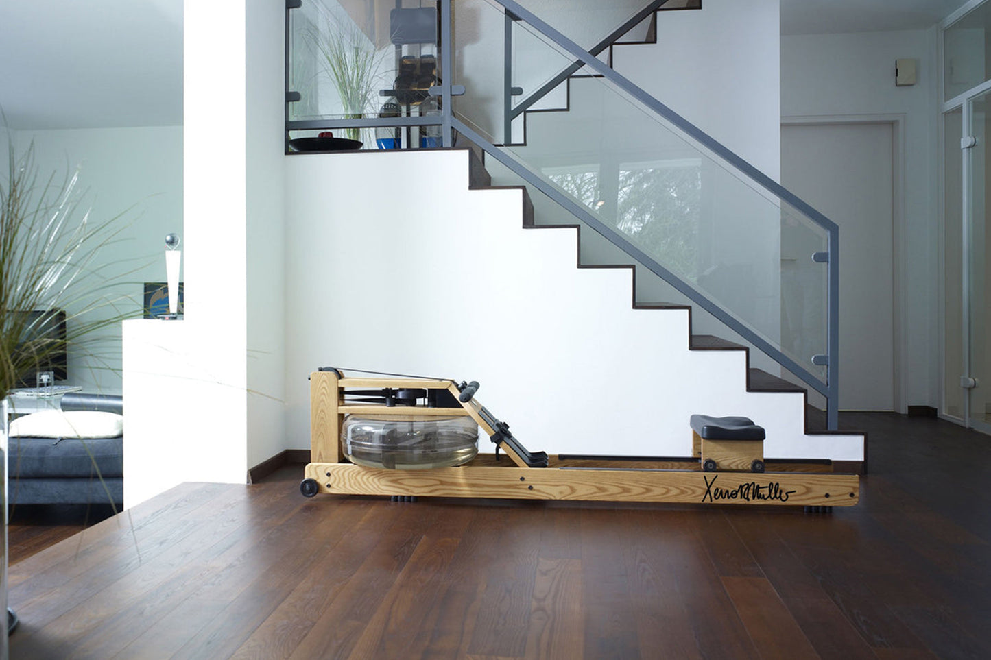 Natural wood rowing machine with glass monitor and black accents on dark wood floor, shown in modern home interior with stair