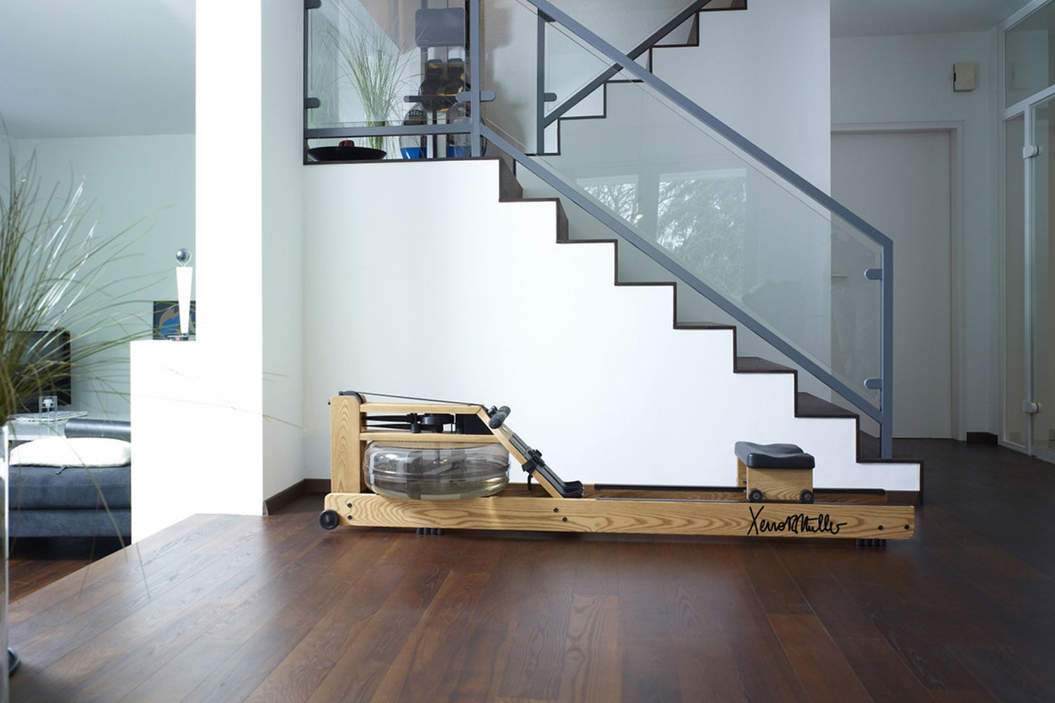 Natural wood rowing machine with glass monitor and black accents on dark wood floor, shown in modern home interior with stair