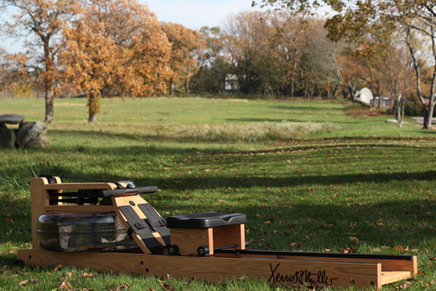WaterRower Xeno Muller Signature Edition rowing machine with tan wood frame and black seat, displayed on grass in autumn park